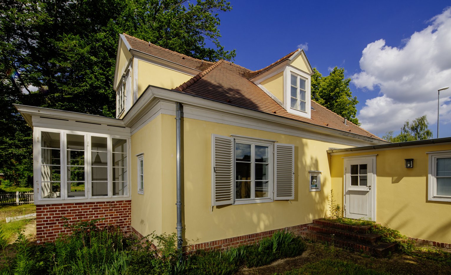 Ein gelbes Haus mit einer Dachgaube, weißen Fensterläden und einem gläsernen Wintergarten.