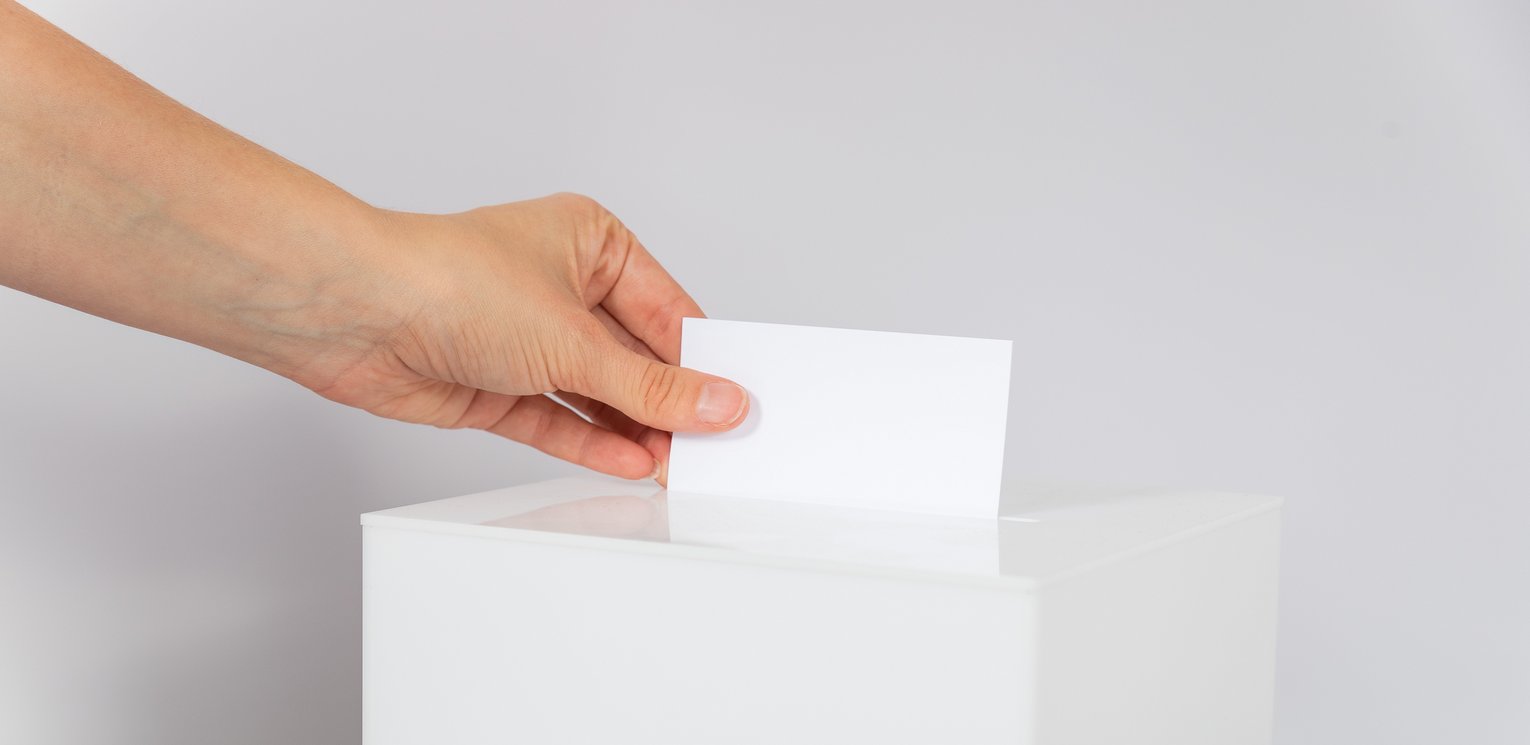 Eine Hand steckt einen weißen Zettel in eine weiße Box mit einem Schlitz.